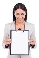 What is there  Curious businesswoman looking at empty paper on a clipboard while standing against white background photo