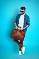 Full length of handsome stylish young man adjusting bag while standing against blue background photo