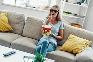 Beautiful young woman watching TV in 3-D glasses and eating popcorn while relaxing on the sofa at home photo