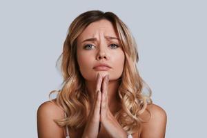 Begging for something. Beautiful young woman looking at camera and keeping hands clasped while standing against grey background photo