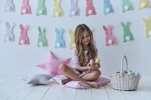 deliciosos dulces de pascua. linda niña sosteniendo huevos de pascua y sonriendo mientras se sienta en la almohada con decoración en el fondo foto