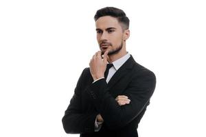 Lost in thoughts. Handsome young man in full suit holding hand on chin and looking away while standing against white background photo