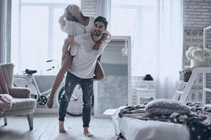 Always happy together. Young couple having fun in bedroom at home while handsome man giving his girlfriend piggy back ride photo