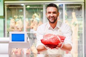 carne de la mejor calidad para ti. joven carnicero alegre estirando un trozo de carne y mirando la cámara mientras está de pie en la caja del supermercado foto