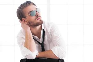 Handsome and confident man. Thoughtful young man in sunglasses holding head in hand and looking away while sitting on the chair photo