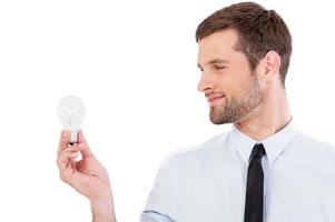 Looking for new ideas. Thoughtful young man in shirt and tie holding lighting bulb and looking at it with smile while standing isolated on white photo