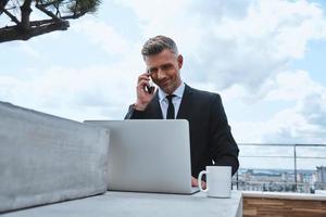 Confident mature man working on laptop and talking on mobile phone while standing on the rooftop terrace photo