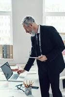 Thoughtful mature man in full suit using laptop and digital tablet while working in modern office photo