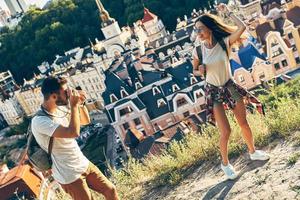 Handsome young man photographing his girlfriend while standing on the top of the hill outdoors photo