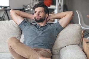 Time to gather all ideas in his head. Handsome young man keeping hands behind his head and looking thoughtful while sitting on the couch at home photo