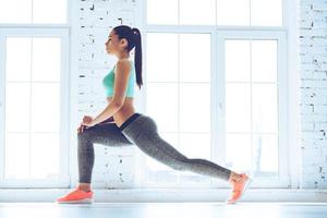 calentamiento previo al entrenamiento. vista lateral de una joven hermosa mujer en ropa deportiva haciendo estiramientos mientras está de pie frente a la ventana en el gimnasio foto