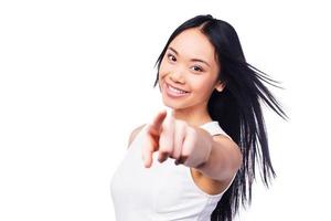 Choosing you Beautiful young Asian woman in pretty dress pointing at camera and smiling while standing against white background photo