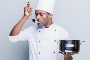 Tasting soup. Confident young African chef in white uniform holding casserole and scoop while standing against grey background photo