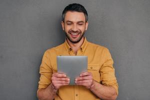 Man with digital tablet. Handsome mature man holding digital tablet and looking at it with smile while standing against grey background photo