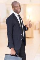 Hombre de negocios exitoso. alegre joven africano con ropa formal sosteniendo un maletín y gesticulando mientras sonríe a la cámara foto