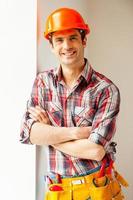 reparador confiado. apuesto joven manitas con casco apoyado en la pared y sonriendo a la cámara foto