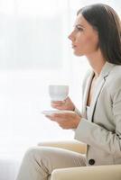 Coffee break. Side view of thoughtful young businesswoman in suit drinking coffee and looking away while sitting at the chair photo