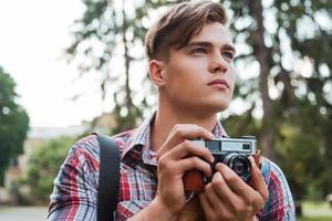 solo inspirado apuesto joven sosteniendo una cámara digital y mirando hacia otro lado mientras está de pie al aire libre foto