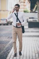 Business talk on a go. Full length of confident young man in glasses holding coffee cup and talking on mobile phone while walking outdoors photo