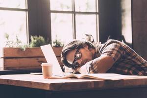 Feeling so tired. Young man in casual wear leaning his head on laptop and sleeping while sitting at his working place in creative office photo