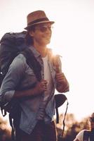 Enjoying early morning hike. Handsome young man in fedora carrying backpack and smiling while walking outdoors photo