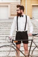 hombre n estilo. un joven barbudo confiado cogido de la mano en su bicicleta y mirando hacia otro lado mientras está de pie al aire libre foto