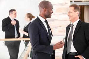comunicacion de negocios. dos hombres de negocios alegres hablando entre ellos mientras sus colegas están de pie en el fondo foto