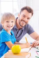 Supporting son in his studying. Side view of cheerful young father helping his son to do homework while both sitting at the table and smiling at camera photo