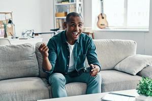 ganando el juego apuesto joven africano gesticulando y sonriendo mientras está sentado en el interior foto