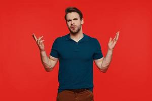 Displeased young man in casual wear making a face and gesturing while standing against red background photo