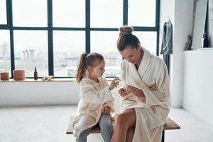 madre e hija en albornoces sonriendo y limpiando dientes mientras hacen la rutina matutina foto