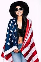 nacido para ser libre. hermosa joven mujer de raza mixta que lleva la bandera americana en los hombros y sonriendo mientras está de pie contra el fondo blanco foto