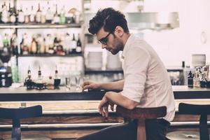 sentirse cansado y con exceso de trabajo. joven frustrado con ropa informal elegante sentado en el mostrador del bar y sosteniendo un vaso foto