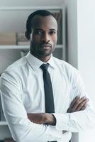 Confident and handsome. Handsome young African man keeping arms crossed and looking at camera while standing in creative office photo