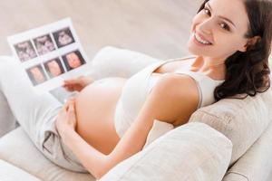 First photo of my baby. Top view of happy pregnant woman sitting on sofa and holding x-ray image of her baby