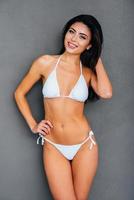 Staying fit and beautiful. Attractive young smiling woman in white bikini holding hand on hip and touching her hair while standing against grey background photo