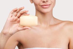 Stay fresh and clean. Cropped image of beautiful young woman wrapped in towel holding a soap bar in her hand while standing against grey background photo