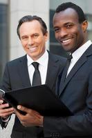 Discussing contract together. Two cheerful business men holding note pad and looking at camera while standing outdoors photo