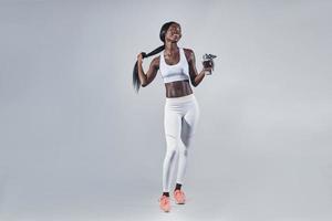 Happy young African woman in sports clothing holding bottle with water and adjusting hair photo