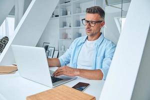 Confident mature man working on laptop while sitting at the office desk photo
