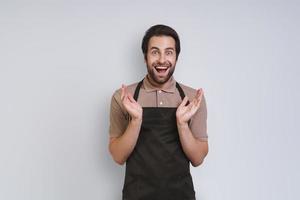 Excited young man in apron looking at camera and gesturing while standing against gray background photo