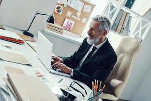 hombre maduro concentrado con traje completo usando una laptop mientras trabaja en una oficina moderna foto