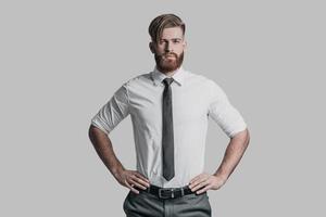 Young and successful. Handsome young man in formalwear keeping hands on hip and looking at camera while standing against grey background photo