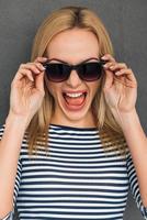 Furious and emotional. Beautiful young woman adjusting her sunglasses and keeping mouth open while standing against grey background photo