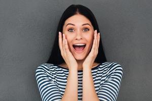 guau, una joven sorprendida tocándose la cara y sonriendo mientras se enfrenta a un fondo gris foto