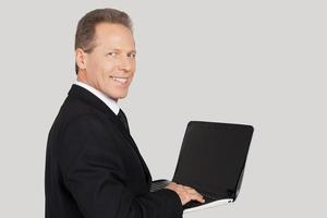 Businessman with laptop. Rear view of senior man in formalwear typing something on laptop and looking over shoulder while standing against grey background photo