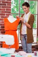 Almost done. Beautiful young fashion designer adjusting dress on the mannequin and smiling with sewing machine on the foreground photo