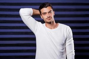 Looking just perfect. Handsome young man in white sweater looking at camera and holding hand behind head while standing against striped background photo