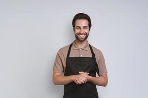 hombre joven guapo en delantal mirando a la cámara y manteniendo las manos juntas mientras está de pie contra el fondo gris foto