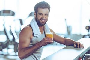 Sip of freshness after great workout. Side view of handsome young men in sportswear holding glass of fresh orange juice and looking at camera with smile while sitting at bar counter at gym photo